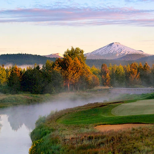 A scenic view of a lush, green golf course next to a reflecting pond, with mist, trees, and a snow-capped mountain in the background ending the sentence.